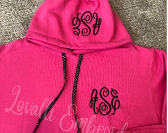 May include: A bright pink hooded sweatshirt featuring black embroidered monogram initials on the chest and hood. The garment includes a front kangaroo pocket and black drawstring cords with a white polka dot pattern for the hood adjustment.