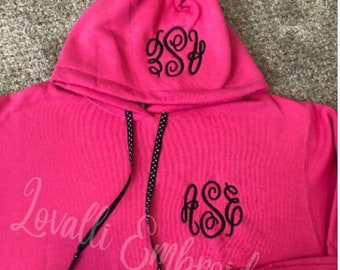May include: A bright pink hooded sweatshirt featuring black embroidered monogram initials on the hood and the left chest. The hoodie includes black drawstring cords with a white polka dot pattern and a large front kangaroo pocket for storage.
