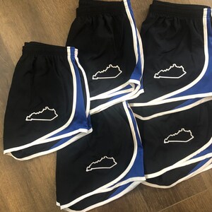 May include: Five pairs of black running shorts with white piping and a blue trim. Each pair has a white outline of the state of Kentucky on the left leg.