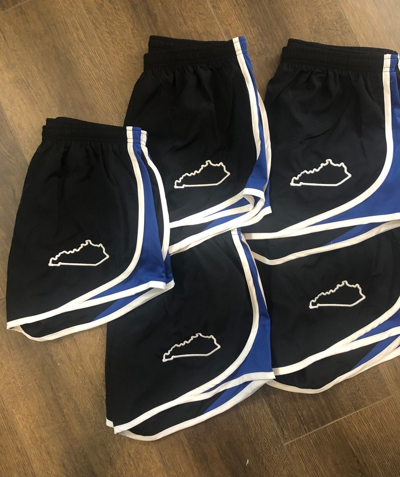 May include: Five pairs of black running shorts with white piping and a blue trim. Each pair has a white outline of the state of Kentucky on the left leg.