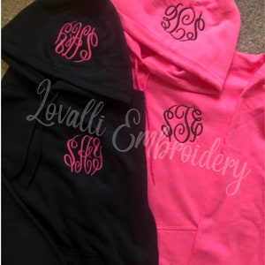May include: A black hoodie with a pink embroidered monogram on the hood and chest, alongside a neon pink hoodie with a black embroidered monogram on the hood and chest. The text Lovalli Embroidery is overlaid across the center of the image.