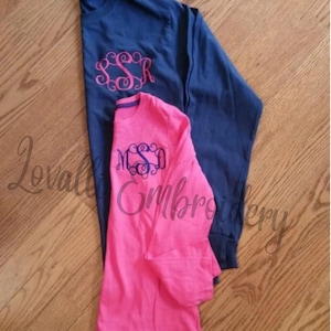May include: Two long-sleeved shirts with embroidered monograms. The top shirt is navy blue with a pink monogram "SSR". The bottom shirt is pink with a blue monogram "MSD".
