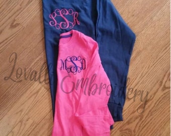 May include: A navy blue long-sleeve shirt with a pink embroidered monogram and a bright pink long-sleeve shirt with a navy blue embroidered monogram. Both shirts feature a crew neckline and long sleeves, with the text Lovalli Embroidery overlaid.