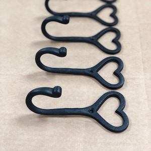 Puede incluir: Un conjunto de cinco ganchos de pared de metal negro dispuestos en una fila vertical. Cada gancho presenta una parte superior curva en forma de J para colgar objetos y una base con forma de contorno de corazón abierto con un orificio.