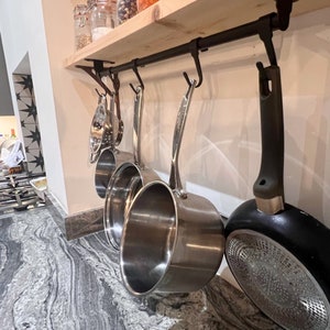 May include: A set of stainless steel pots and a black frying pan hanging from a black metal rack mounted on a wooden shelf. The pots and pan are hanging from hooks. The pots are all different sizes. The frying pan has a black handle and a silver bottom.