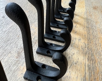 Könnte beinhalten: Eine Reihe von Wandhaken aus schwarzem Gusseisen. Jeder Haken hat ein geschwungenes, texturiertes Design mit einem abgerundeten Oberteil und einem kleineren unteren Haken. Die Basis ist rechteckig und verfügt über zwei vorgebohrte Montagelöcher.