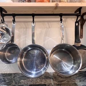 May include: A black metal kitchen rail mounted under a wooden shelf with five black hooks. Hanging from the hooks are a stainless steel colander, two stainless steel saucepans, a stainless steel frying pan, and a black non-stick frying pan.