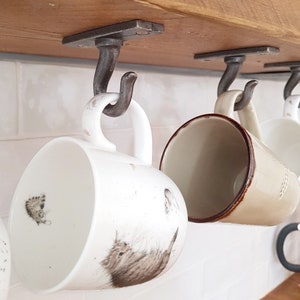 May include: Four rustic metal hooks mounted on a wooden shelf, holding a variety of mugs. The mugs are white with brown accents and feature a cat design.