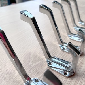 Puede incluir: Una fila de seis ganchos de metal plateado, dispuestos en un patrón escalonado sobre una superficie de madera clara. Los ganchos son todos del mismo tamaño y forma, con un diseño ligeramente curvado.
