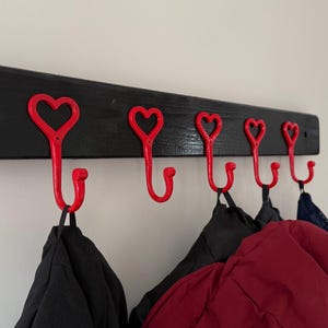 May include: A black wooden coat rack with six red heart-shaped hooks. Several coats hang from the hooks. The rack is mounted on a white wall.