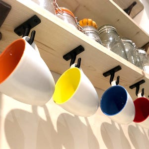 Può includere: Cinque tazze in ceramica appese a ganci su uno scaffale di legno. Le tazze sono bianche con accenti colorati: arancione, giallo, blu, rosso e verde.