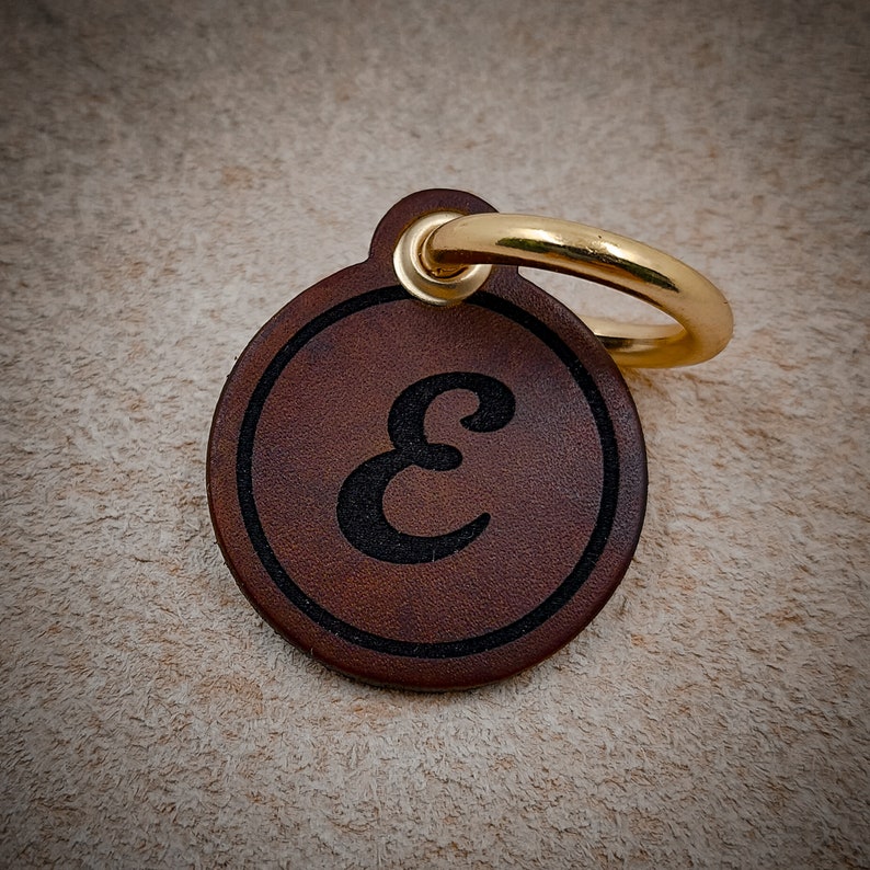 Op de afbeelding: Een ronde bruine leren hanger met een zwarte, in reli&euml;f aangebrachte letter E in het midden, omsloten door een cirkelvormige rand. Een glanzende goudkleurige metalen ring is bevestigd door een klein lipje aan de bovenkant van de schijf.