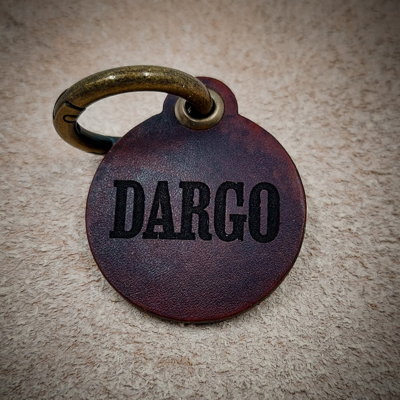 Op de afbeelding: Een ronde donkerbruine leren naamplaat voor huisdieren met de naam DARGO in zwarte hoofdletters. De plaat is voorzien van een bronzen metalen ring die door een verstevigd oogje aan de bovenkant van de leren schijf is bevestigd.