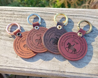 Op de afbeelding: Vier ronde leren sleutelhangers met metalen ringen. Elke hanger toont een silhouet van een paard met ruiter, een naam en een cirkelvormige tekst. De tekst luidt: Make the wrong things easy, Wish, In order to fly, Dargo, Ride forward with joyful confidence.