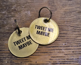 Op de afbeelding: Twee ronde, messingkleurige metalen labels, elk voorzien van een kleine ring aan de bovenkant. De labels zijn gegraveerd met de zwarte tekst TWEET ME MAYBE. Het oppervlak van het metaal heeft een geborstelde afwerking en ze overlappen.