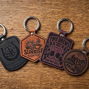 May include: Four leather keychains with metal rings. One black shield-shaped with 'LSTC LEATHER', one tan hexagon with 'SUNRISE COFFEE', one rectangular burgundy with 'TWO BREWS BAR AND GRILL', and one round brown with 'PLEASANT PRAIRIE RACING' text.