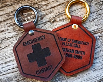 Op de afbeelding: Twee zeshoekige bruine leren labels met metalen ringen. Eén label heeft een zwart kruis en de tekst EMERGENCY CONTACT. Het andere label toont de tekst IN CASE OF EMERGENCY PLEASE CALL: NATHAN SMITH 555-888-8888. Beide labels hebben een metalen klinknagel.