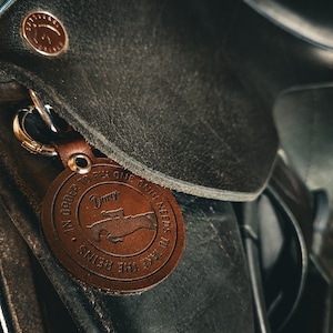 Puede incluir: Un llavero de cuero marrón con un cierre plateado, sujeto a una silla de montar de cuero negro. El llavero tiene una etiqueta circular con el texto "In order to fly one only needs to take the reins." y un gráfico de una mano.