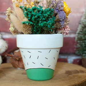 Peut inclure: Un pot en terre cuite blanc avec un motif géométrique vert et noir, rempli d'un bouquet de fleurs séchées, comprenant des fleurs violettes, jaunes et blanches.