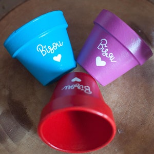 Peut inclure: Trois petits pots de fleurs peints à la main en bleu, violet et rouge. Chaque pot porte le mot "Bisou" et un cœur peint en blanc. Les pots sont disposés sur une surface en bois.