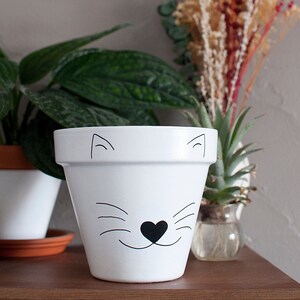 Peut inclure: Pot en céramique blanche avec un visage de chat noir peint sur le devant. Le chat a un nez en forme de cœur et des moustaches.