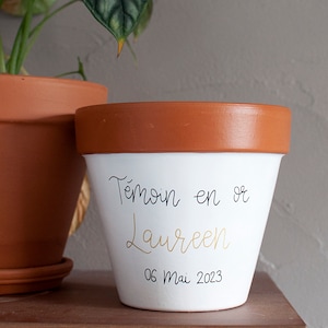 Peut inclure: Pot de fleurs en terre cuite blanche avec un bord brun. Le pot est décoré d'une inscription noire qui dit "Temoin en or" et "Laureen" en lettres dorées. La date "06 Mai 2023" est écrite en noir sous le nom.
