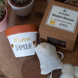 Peut inclure: Un pot de fleurs en céramique blanc et orange avec l'inscription "Maman D'AMOUR" en lettres dorées et deux cœurs dorés. Une boîte en carton avec l'inscription "Kit A Planter Mission Banane" et un petit sac sont également présents.
