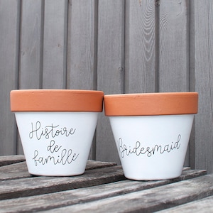 Peut inclure: Deux pots de fleurs en céramique blanche avec un bord en terre cuite. L'un porte l'inscription "Histoire de famille" en écriture noire, l'autre "Bridesmaid". Les pots sont sur une surface en bois.