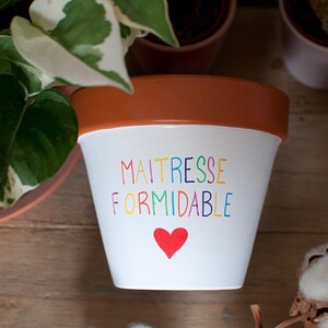Peut inclure: Un pot de fleurs en terre cuite blanc avec un rebord marron. L'avant présente le texte multicolore "MAITRESSE FORMIDABLE" écrit dans un style manuscrit, positionné au-dessus d'une petite icône de cœur rouge solide centrée sur la surface blanche.