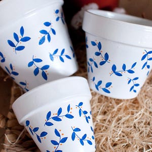 Peut inclure: Trois pots de fleurs en céramique blanche avec un motif de feuilles bleues et des points orange. Les pots sont disposés sur une surface tissée, présentant un motif floral décoratif. Les pots sont de tailles variables.