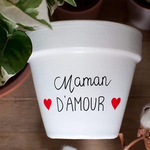Peut inclure: Pot de fleur en céramique blanche avec l'inscription "Maman D'AMOUR" en écriture noire, encadrée de deux cœurs rouges. Un cadeau parfait pour la fête des mères. Idéal pour les plantes.