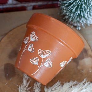 Peut inclure: Pot de fleurs en terre cuite avec un motif floral blanc. Le pot est de couleur brun clair et a une finition lisse et mate.