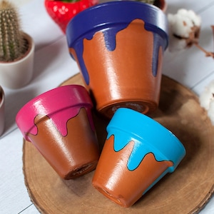 Peut inclure: Trois petits pots de fleurs en terre cuite peints à la main. Chaque pot est marron avec une couleur contrastante dégoulinant du bord : bleu, rose et bleu clair. Les pots sont disposés sur une tranche de bois, avec d'autres petites plantes en arrière-plan.