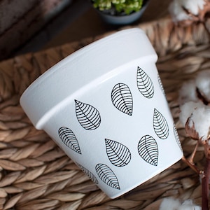 Peut inclure: Pot en céramique blanche avec un motif de feuilles noires. Le pot est posé sur une surface en osier tressé.