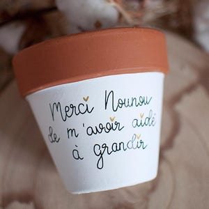 Peut inclure: Petit pot de fleur en céramique blanche avec un bord en terre cuite. Le pot porte l'inscription manuscrite noire « Merci Nounou de m'avoir aidé à grandir » avec de petits cœurs dorés. Idéal pour les petites plantes.