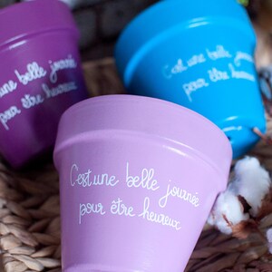 Peut inclure: Trois pots de fleurs en terre cuite peints avec des peintures de différentes couleurs et des lettres blanches. Les pots sont sur une surface en osier tressé. Le texte sur les pots est "C'est une belle journée pour être heureux".