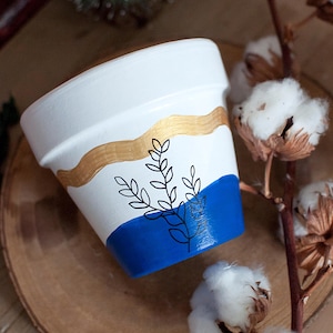Peut inclure: Un pot en terre cuite blanc avec un motif peint bleu et or. Le pot présente un motif floral noir stylisé.