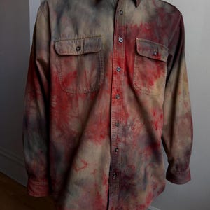 May include: A long-sleeve button-down shirt featuring a mottled tie-dye pattern in shades of deep red, charcoal gray, and muted brown. The shirt includes a standard collar, a front button placket, and two chest pockets with button-down flaps.