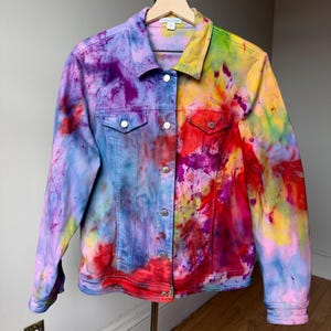 May include: A long-sleeve denim jacket featuring a vibrant, multi-colored tie-dye pattern with splashes of purple, blue, yellow, red, and pink. The jacket includes a classic collar, a button-down front with silver-toned metal buttons, and two chest pockets.