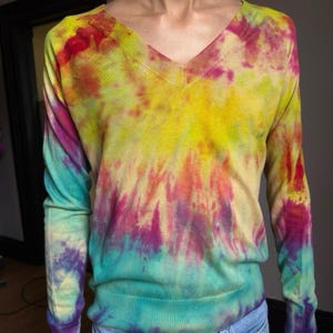May include: A long-sleeved V-neck sweater with a vibrant tie-dye pattern. The colors include yellow, red, purple, and teal, creating a unique and eye-catching design. The sweater is made of a soft material and has a classic fit.