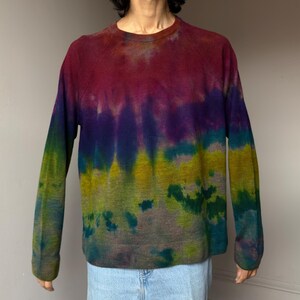 May include: A long-sleeved sweater with a tie-dye pattern. The colors transition from red at the top to purple, blue, yellow, and green towards the bottom. The sweater has a crew neck and raglan sleeves. The garment is made of a soft, knit material.
