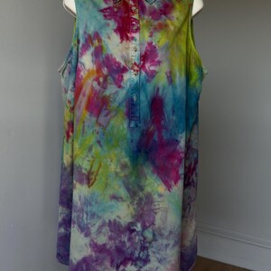 May include: A sleeveless collared shirt featuring a vibrant tie-dye pattern in shades of magenta, bright blue, lime green, and deep purple. The garment has a front button placket with small metallic buttons extending from the collar to the chest.