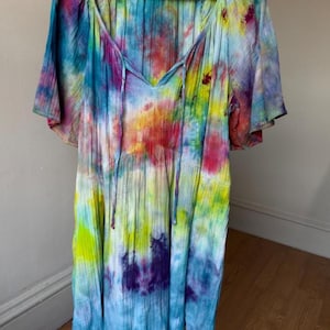 May include: A short-sleeved dress featuring a vibrant tie-dye pattern in shades of blue, yellow, red, purple, and green. The garment has a V-neckline with thin fabric ties, a gathered bodice, and a textured, crinkled fabric finish throughout.