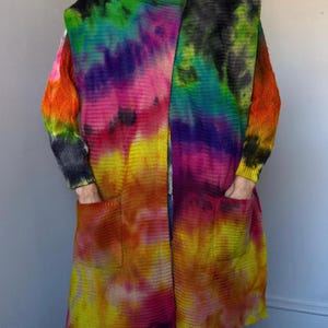 May include: A long, open-front cardigan in a vibrant tie-dye pattern. The color palette includes shades of yellow, orange, pink, purple, green, and black. The cardigan features long sleeves, pockets, and a unique asymmetrical design. The garment is made of a knitted material.