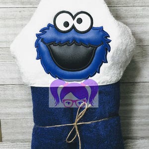 May include: A hooded towel featuring a Cookie Monster design. The hood is white with a blue and black Cookie Monster face. The towel is a dark blue color and tied with twine.