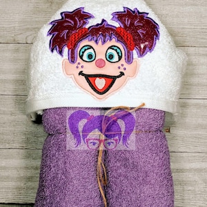May include: A white hooded towel with an embroidered cartoon face and purple hair. The towel is wrapped in a matching purple towel and tied with twine. The cartoon face has blue eyes, a red mouth, and a pink nose.