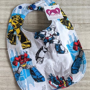 May include: White baby bib with a blue trim and snap closure. The bib features a repeating pattern of colorful Transformers characters in action poses, with a blue and white cityscape background.