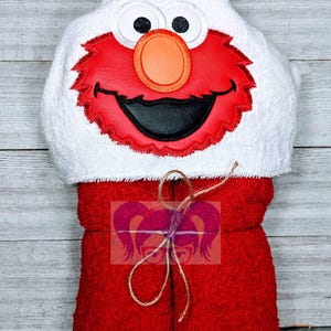 May include: A red and white hooded baby towel featuring a cartoon character face. The hood has large white eyes, a round orange nose, and a red mouth. The towel is tied with a natural twine.