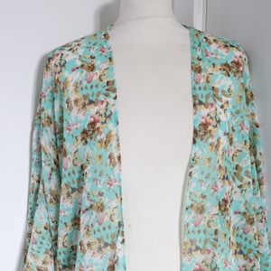 May include: A sheer, open-front kimono in a light turquoise color, adorned with a floral pattern in shades of brown, pink, and white. The kimono is draped over a white mannequin, showcasing its flowing design and lightweight fabric.