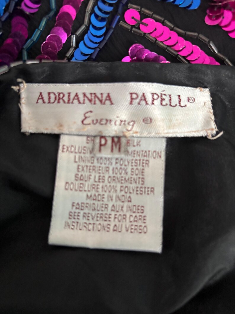 Puede incluir: Primer plano de una prenda de noche negra con lentejuelas y abalorios rosas y azules. La etiqueta dice "ADRIANNA PAPELL Evening". El forro es 100% poli&eacute;ster y el exterior es 100% seda.